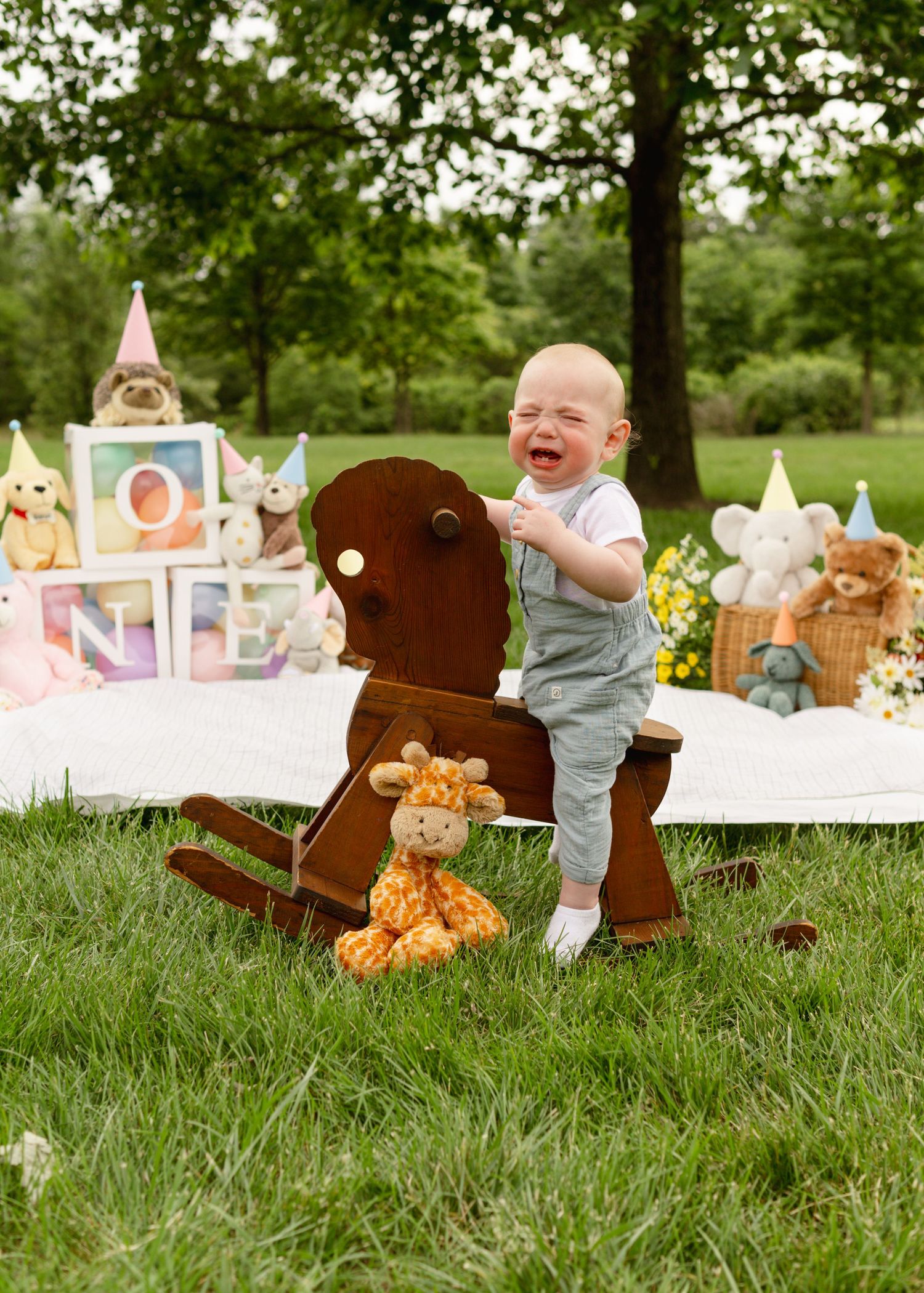 A baby plays in grassy outdoor setting with teddy bears and decorative party banners at a garden party photoshoot.