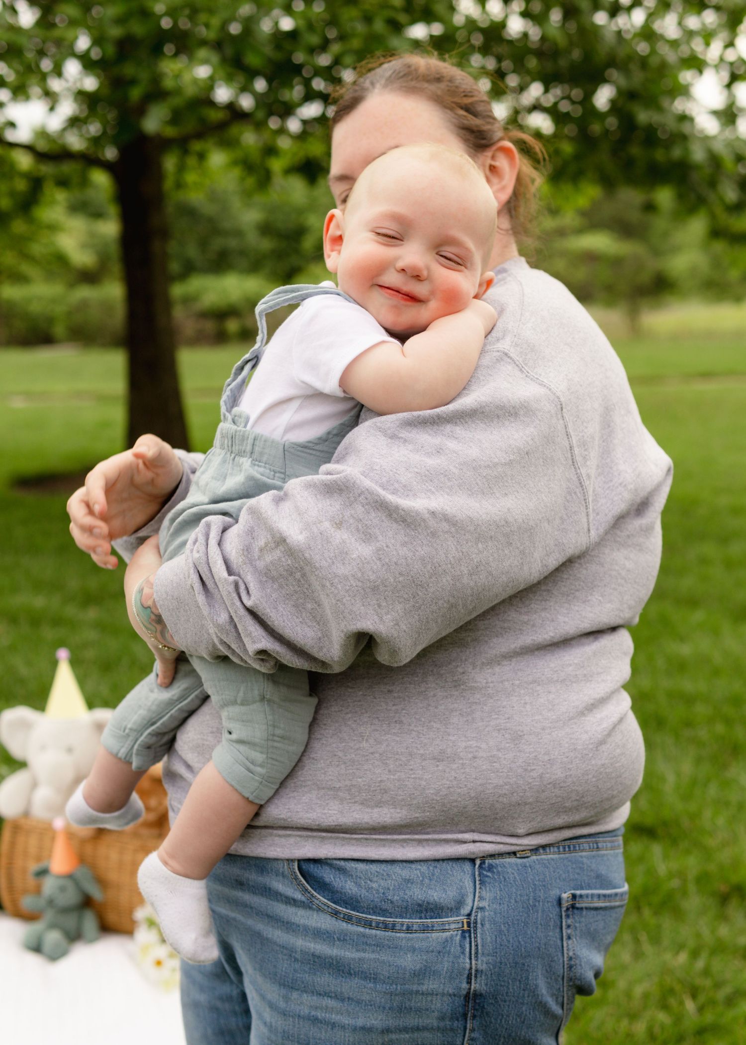 A tender embrace captured outdoors on a spring day with green trees and a picnic basket in the background.