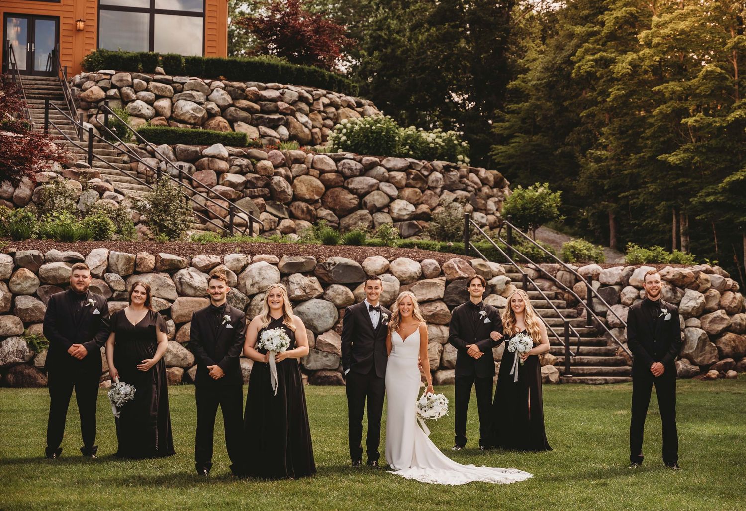 Wedding party lined up against stone wall backdrop in elegant formal attire.