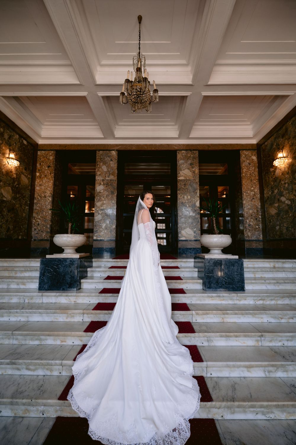 A bride in an elegant white lace wedding gown with long train stands on red-carpeted marble stairs in a grand hall.