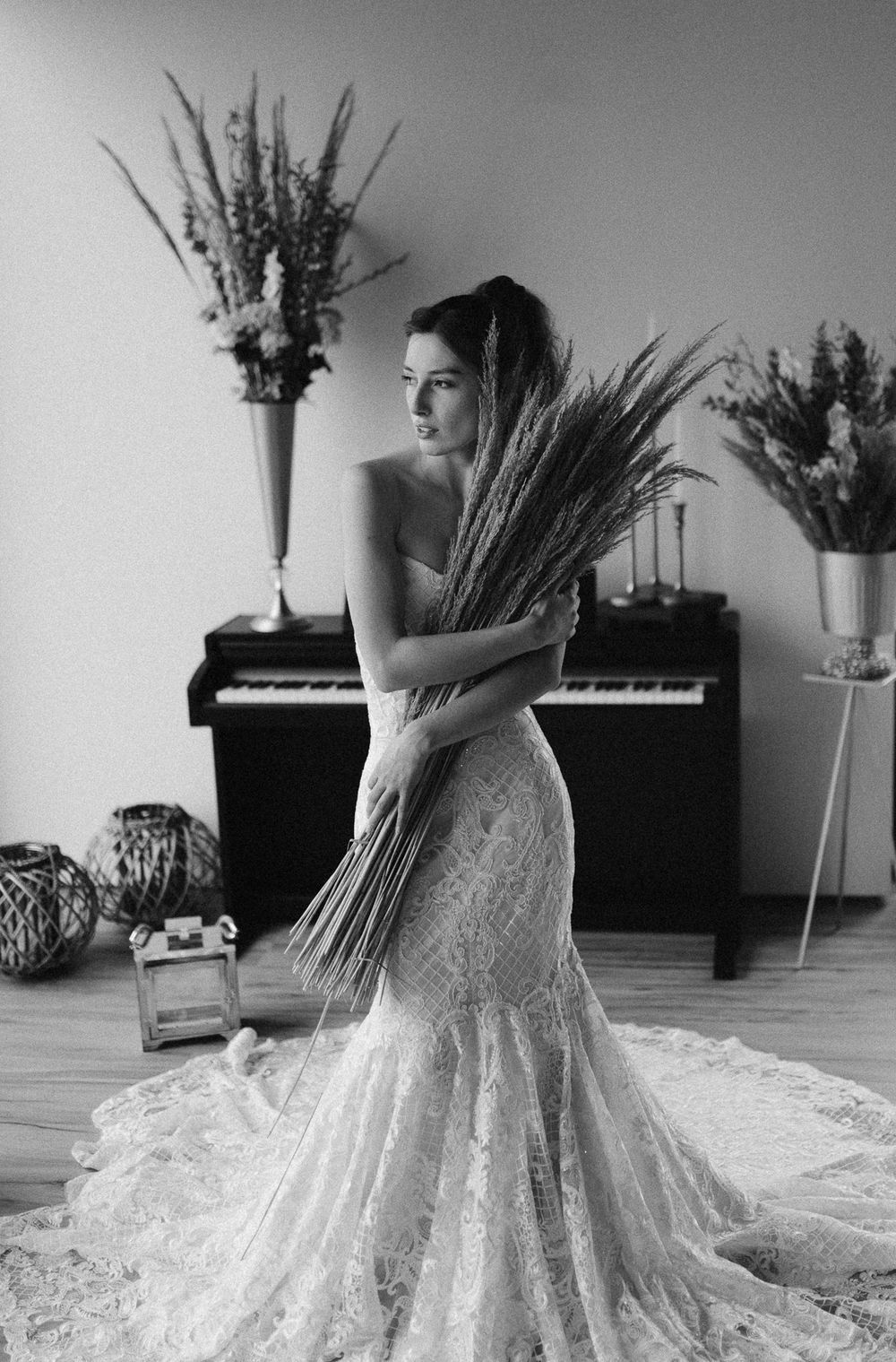 Black and white artistic portrait in elegant lace wedding dress holding wheat stalks against piano and floral decor.