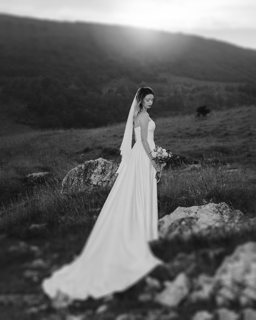A bride in a flowing white dress stands on a rocky hillside in dramatic black and white photography.