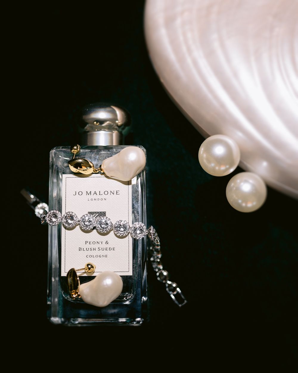 A Jo Malone perfume bottle styled with pearls and a crystal bracelet against a dark background.