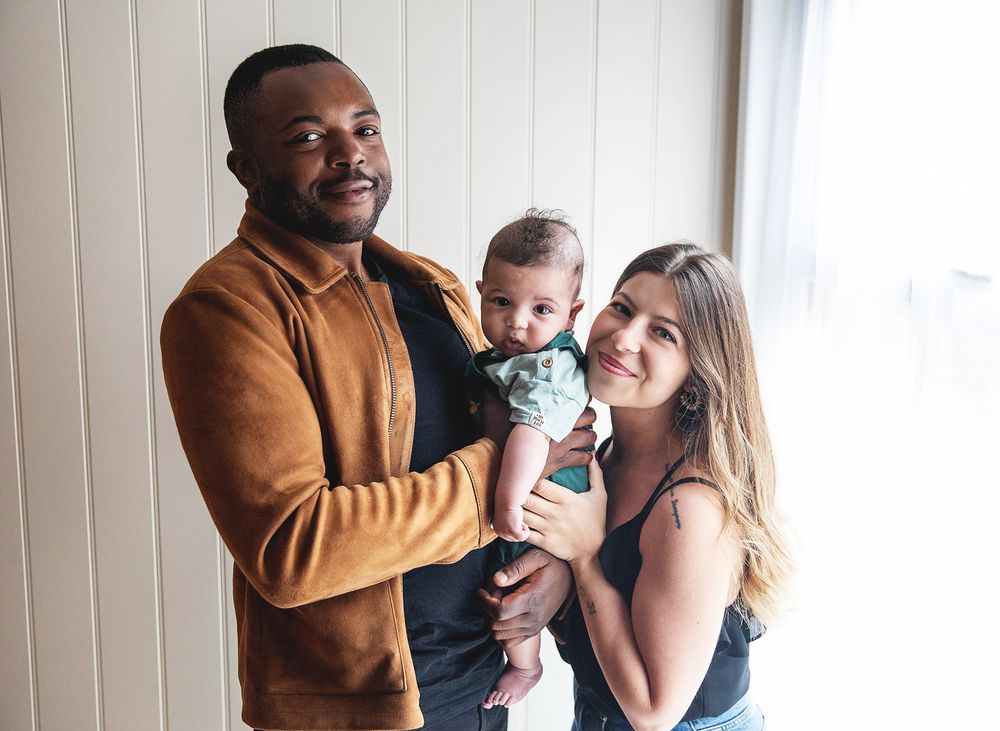 Happy couple holds their baby together while standing by a window in their home. During a family photography session in Sidcup