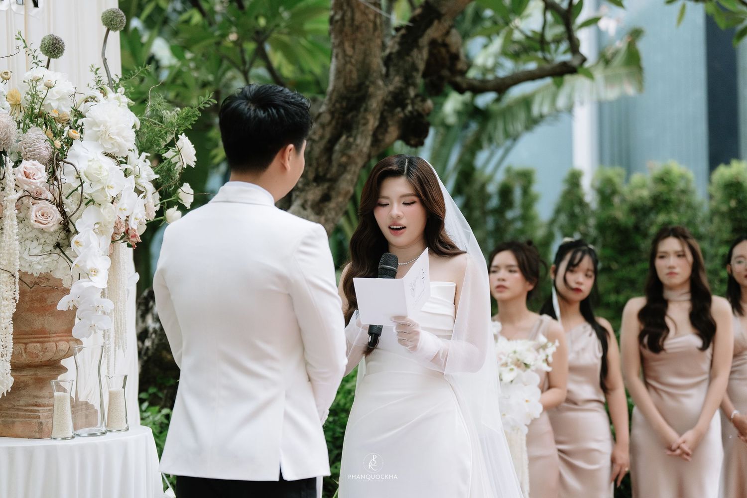 Bride reads wedding vows to groom at outdoor ceremony with bridesmaids standing behind in champagne dresses.
