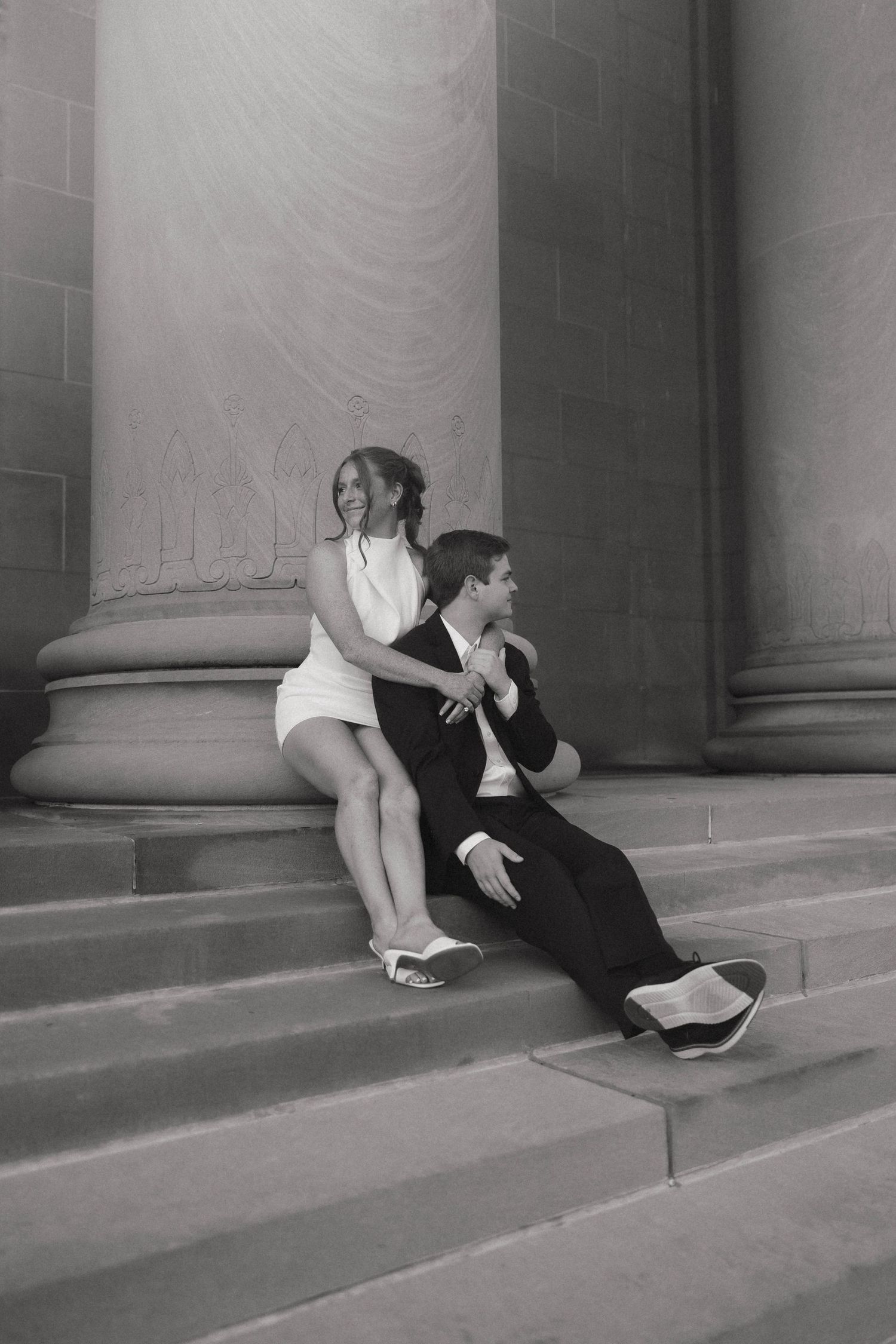 Classic black and white photo of a couple seated on grand stone steps, woman in white dress and man in dark suit.