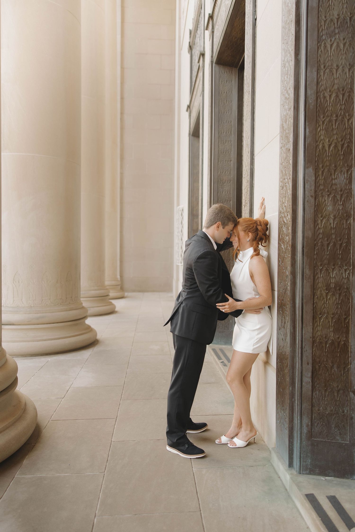A couple shares an intimate moment in a hallway with elegant columns, the bride in a short white dress and groom in dark suit.