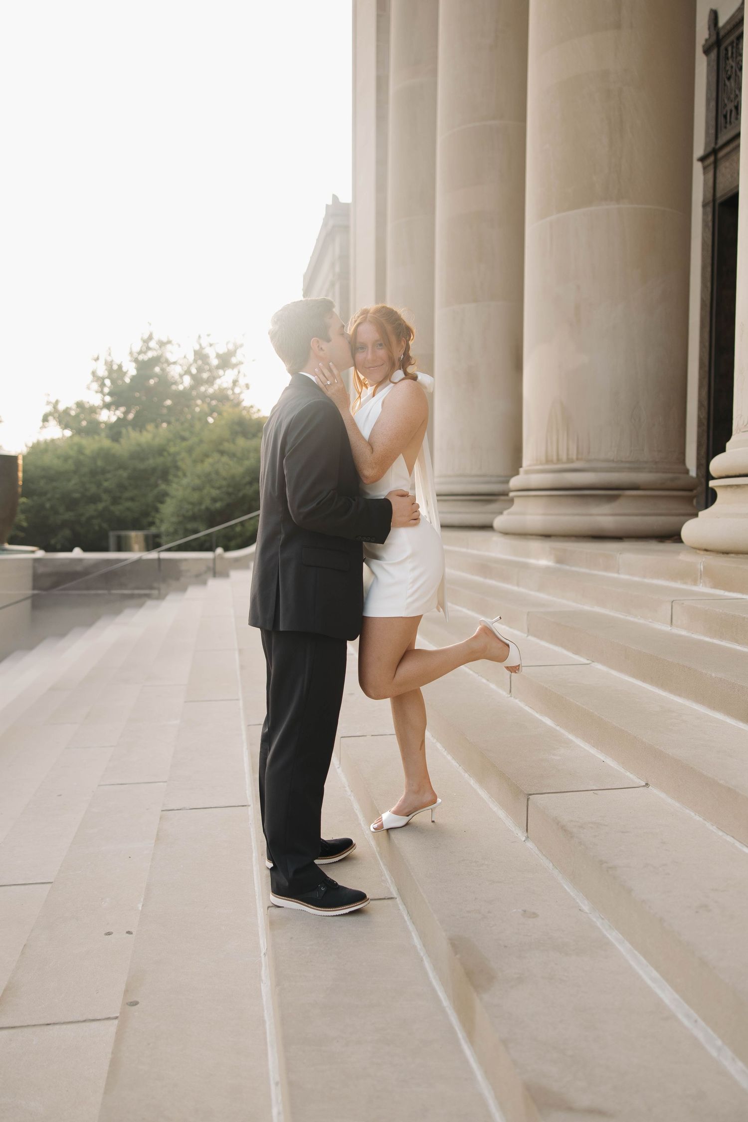 A romantic couple in formal attire embraces on stone steps at sunset near classical columns.