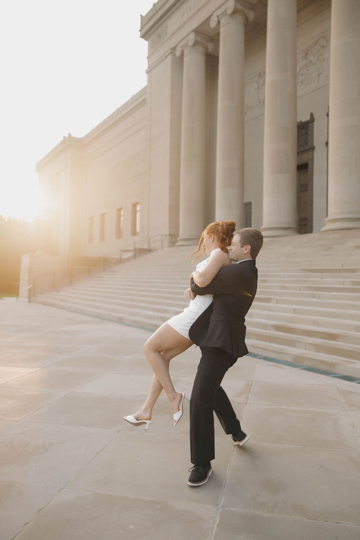 A couple embraces in a romantic dance pose on the steps of a grand building with classical columns at sunset.