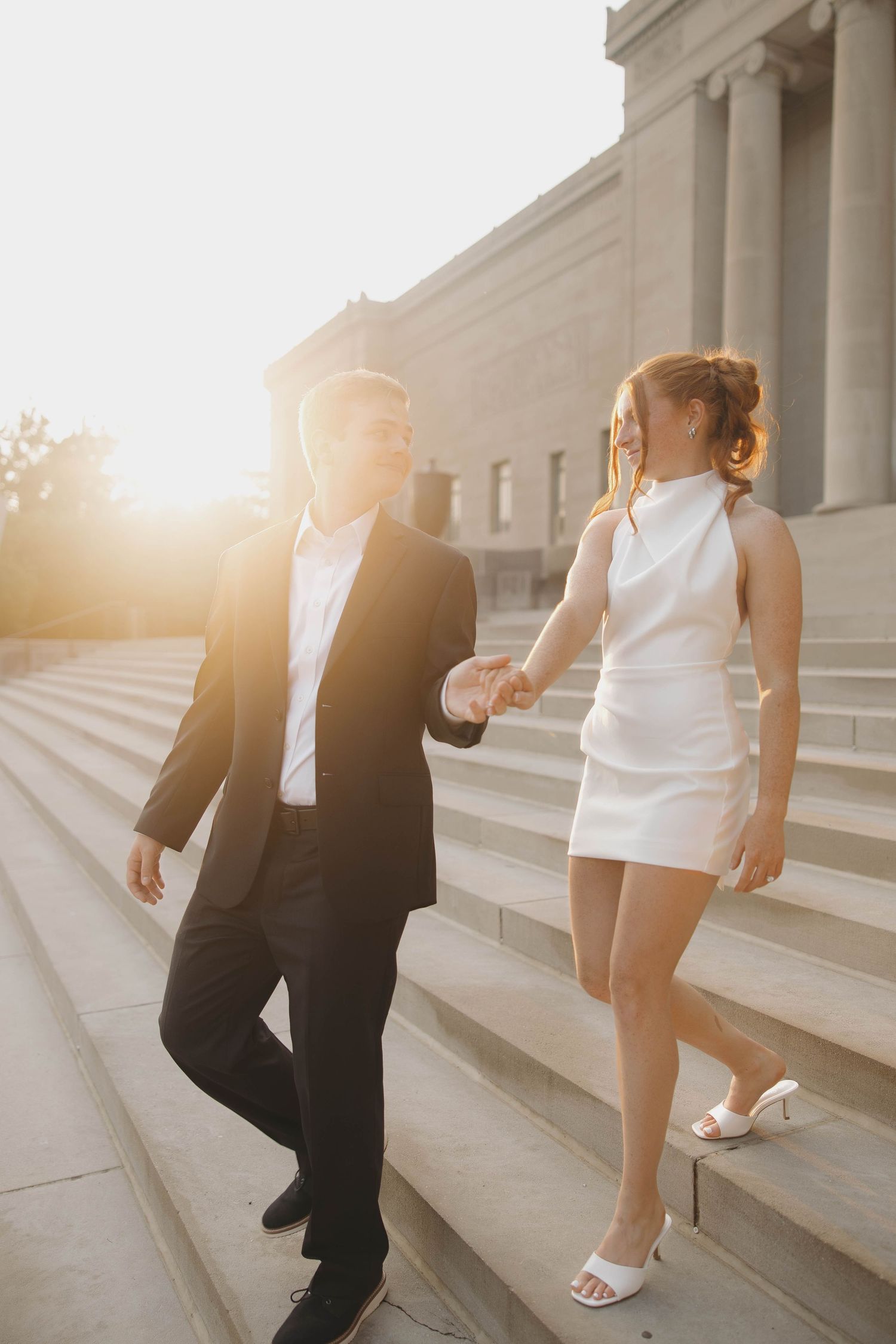 A couple descends marble steps at golden hour, with a man in a black suit and woman in a white dress walking elegantly.