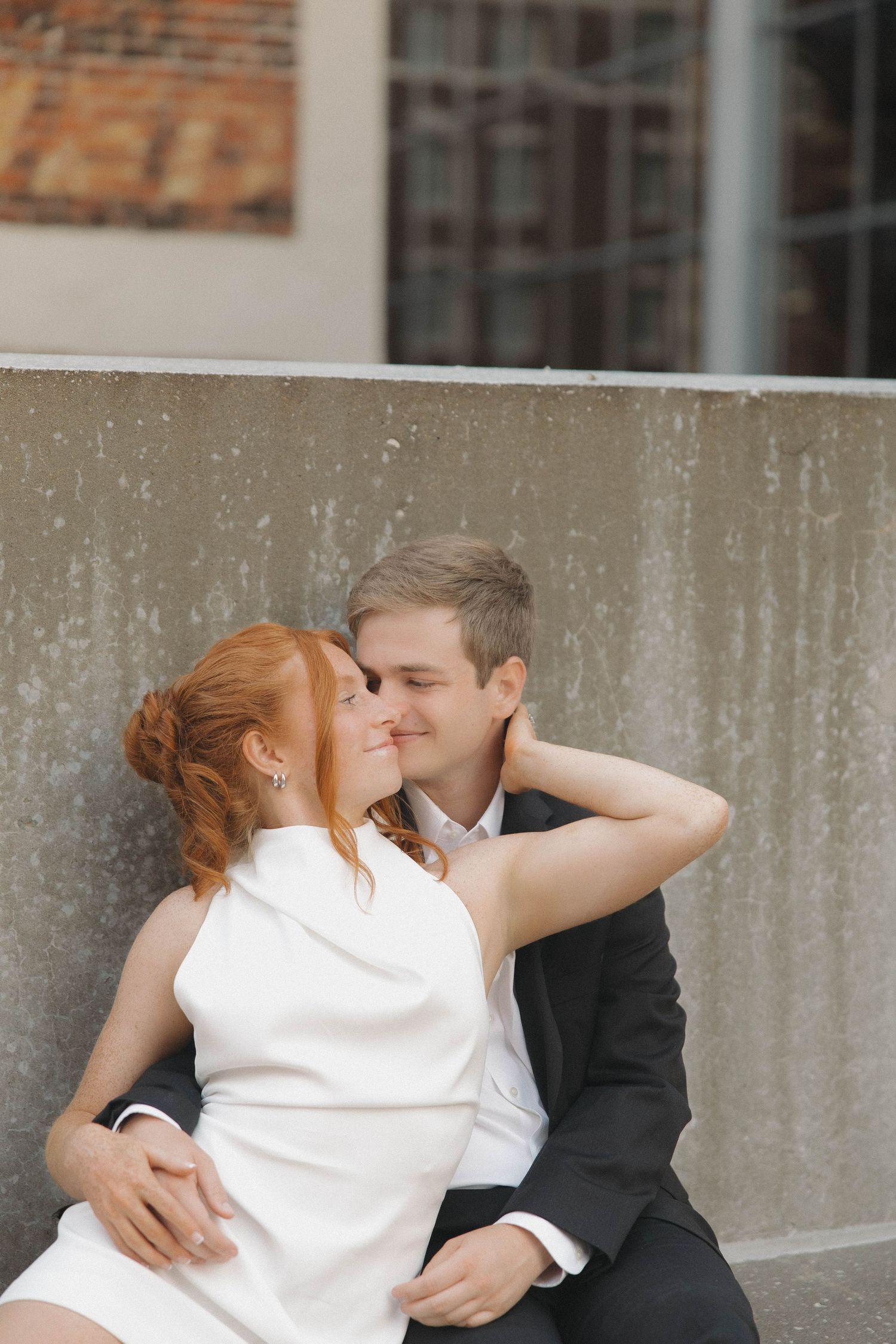 A tender romantic moment between a couple in wedding attire sitting against a concrete wall.