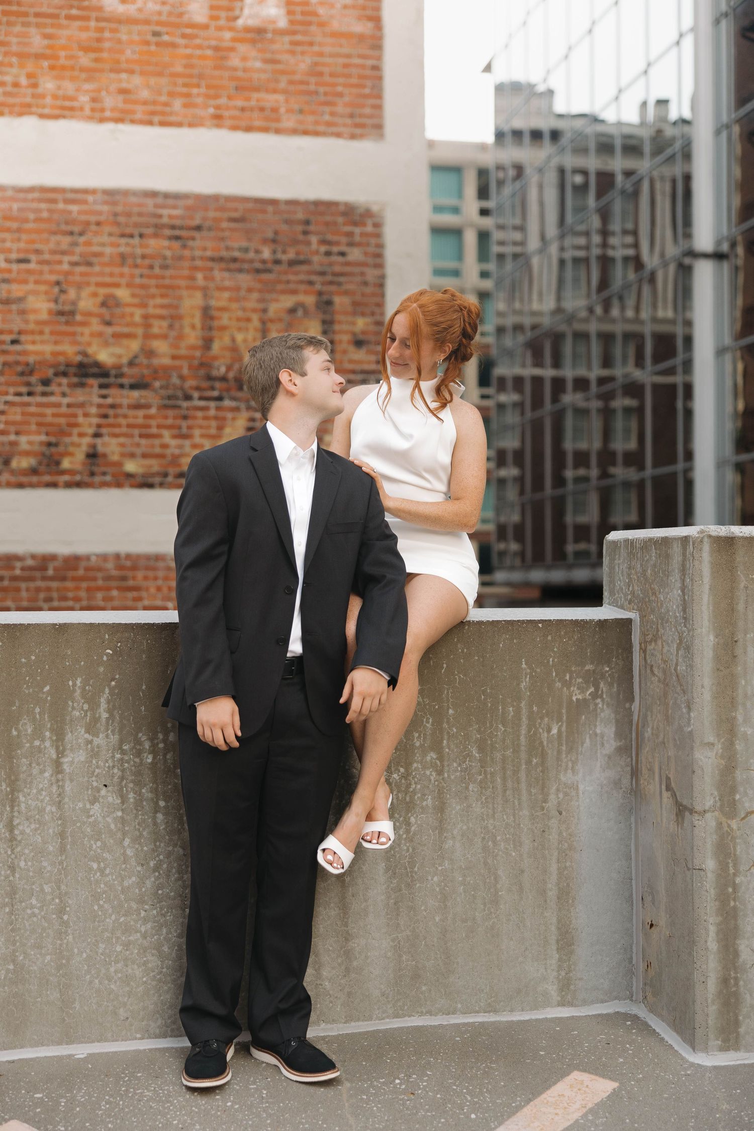 A couple in formal attire interacts playfully by a brick wall in an urban setting.