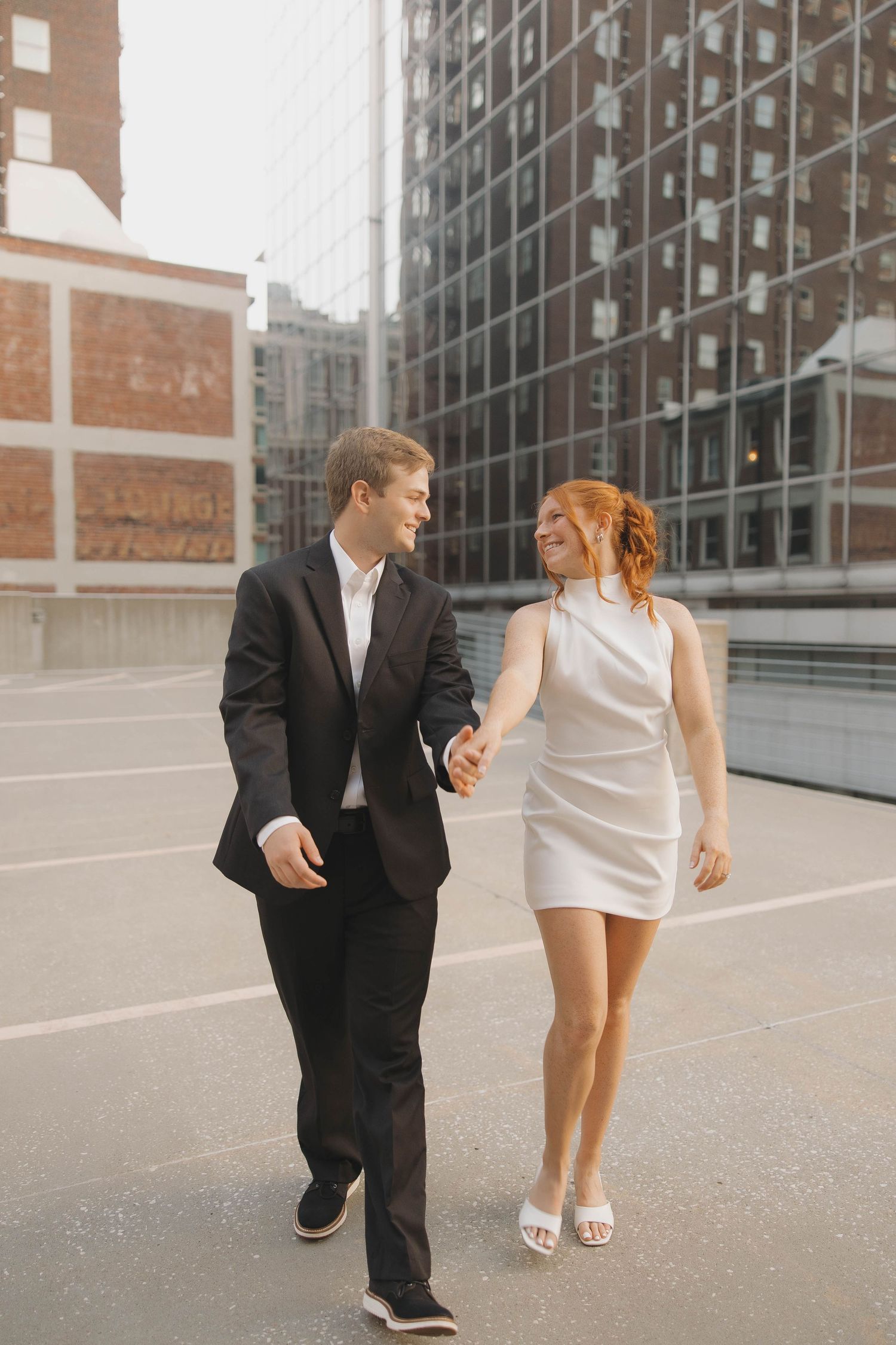 Modern wedding couple with groom in black suit and bride in short white dress posing against urban architecture.