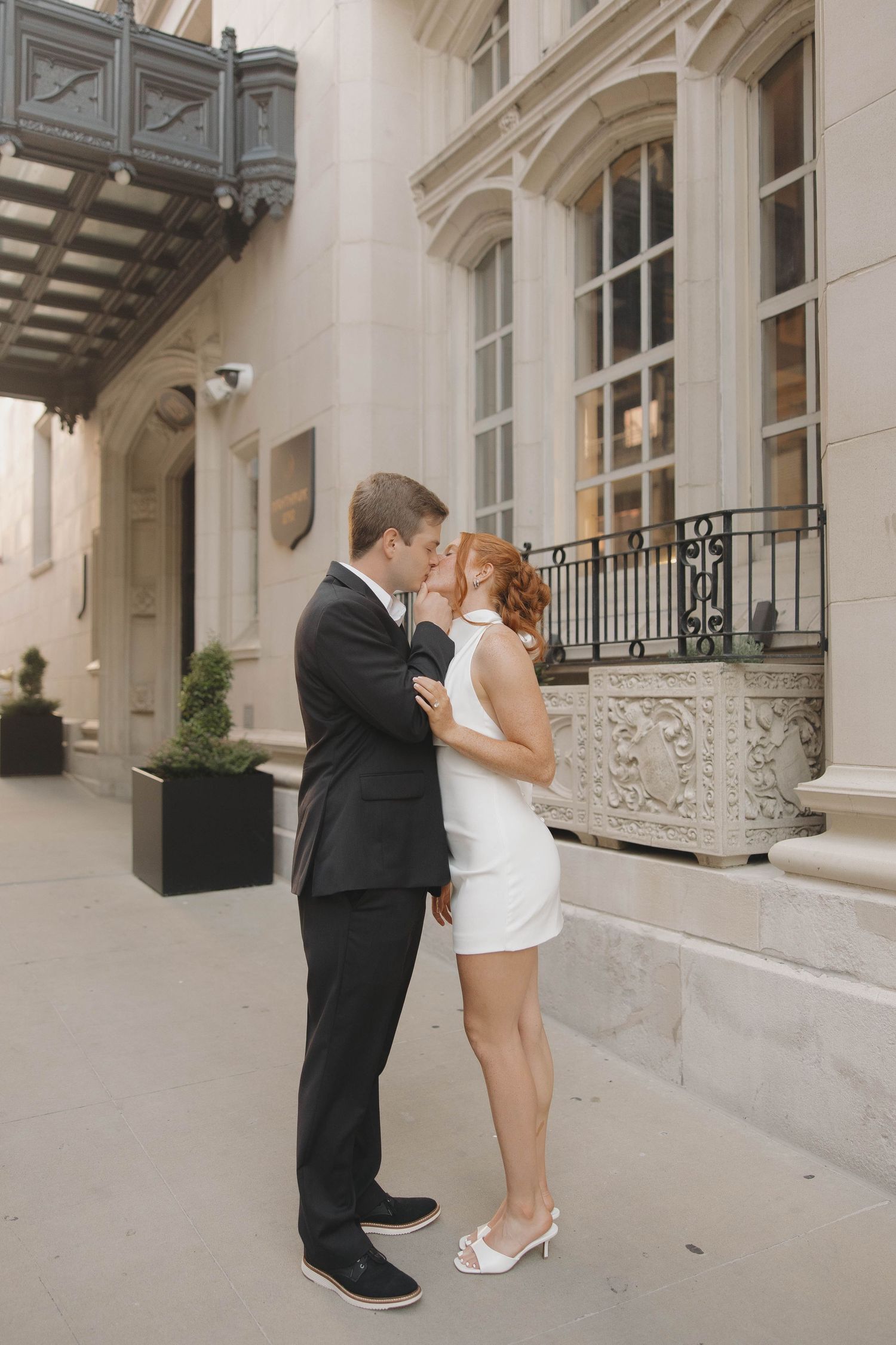 A couple shares a romantic moment outside an elegant building with ornate architecture and a decorative balcony.