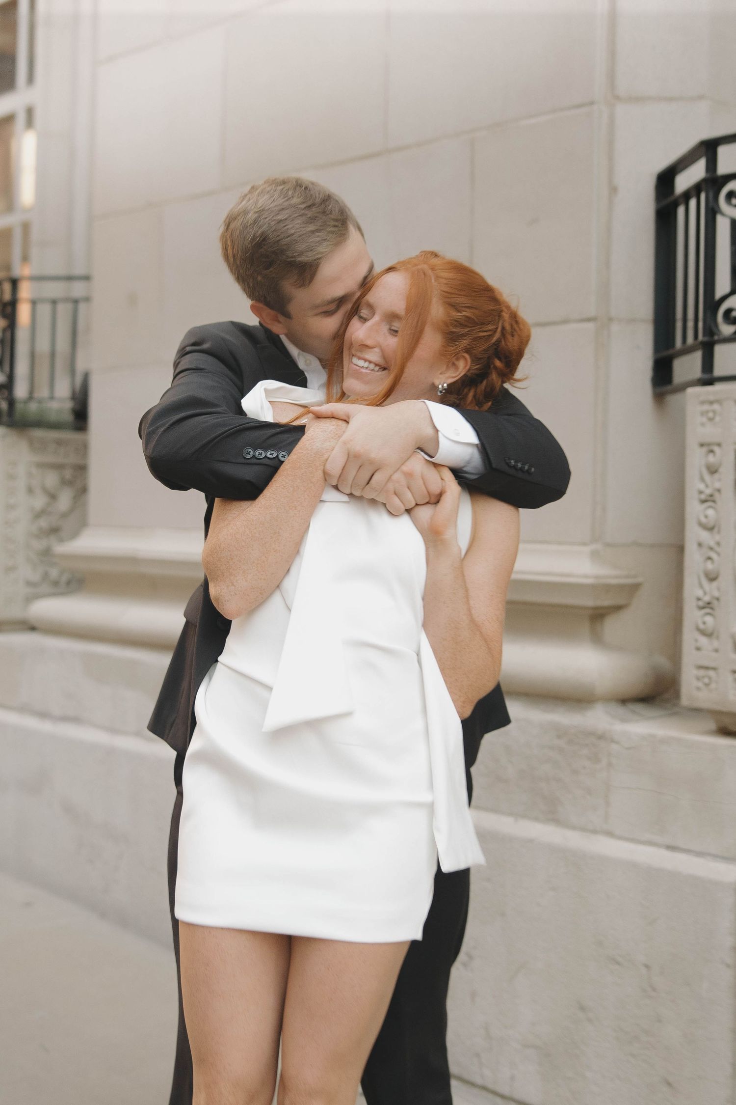 A couple embraces outdoors, with the bride wearing a short white dress and groom in classic black suit.