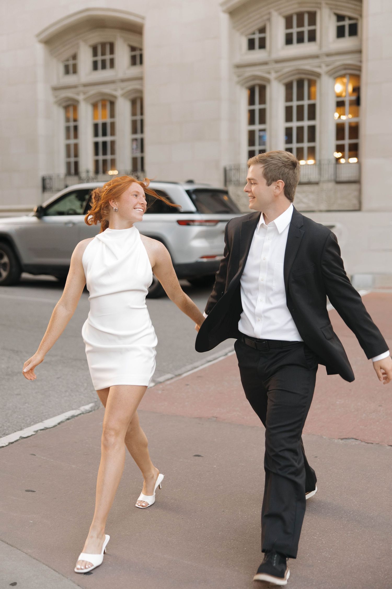 A playful couple dancing on a city sidewalk in formal attire with classic architecture behind them.