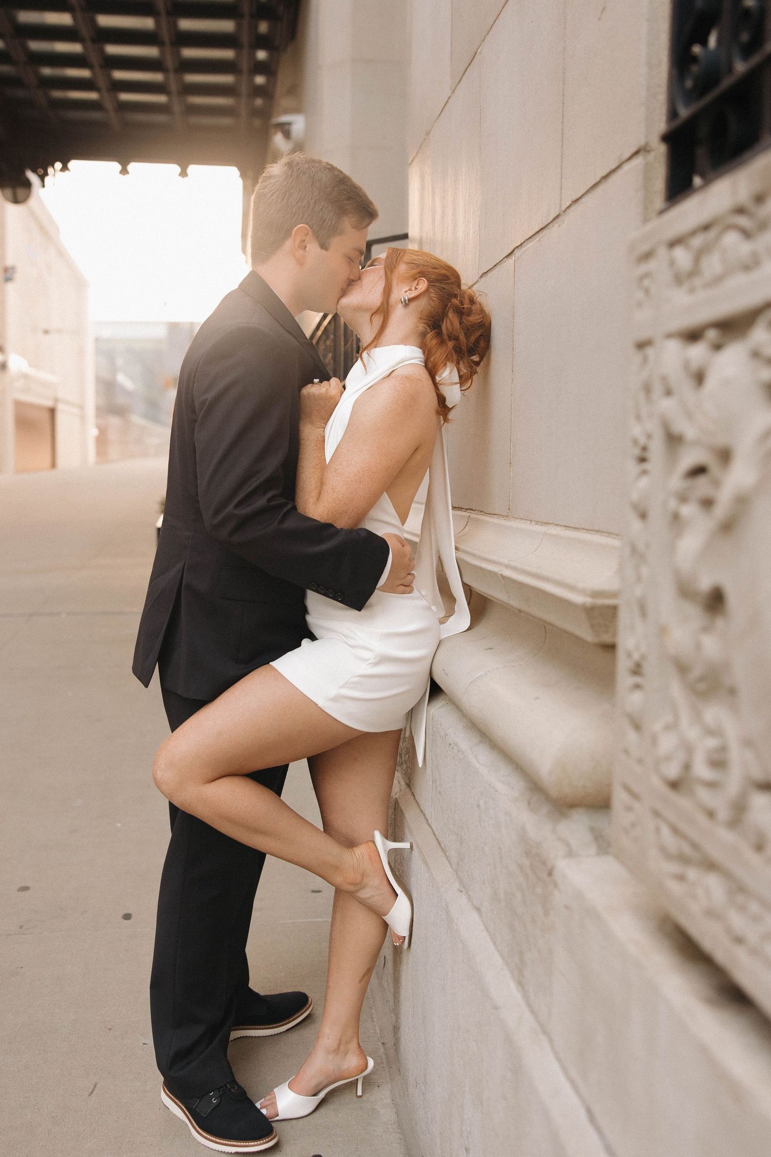 A romantic couple in wedding attire share an intimate moment against a stone wall in a vintage-toned photograph.