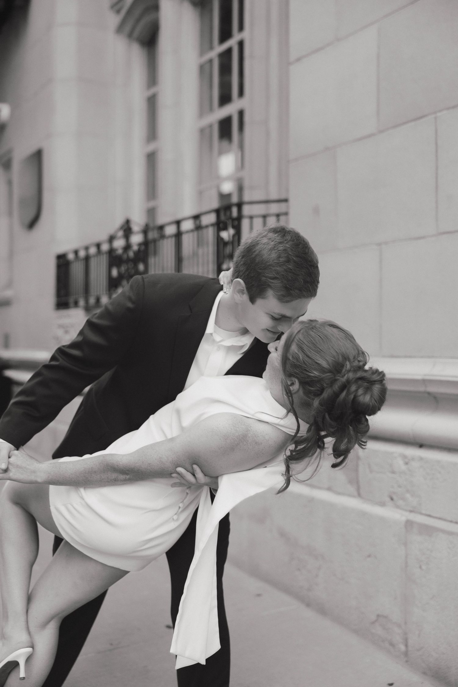 A couple in vintage attire share a romantic kiss while leaning over a marble railing in a black and white photograph.