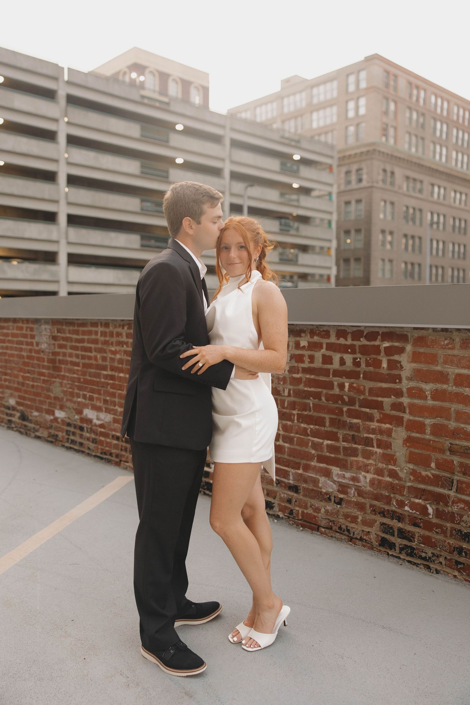 A couple in formal attire embrace on a rooftop parking deck with brick walls and city buildings in the background.