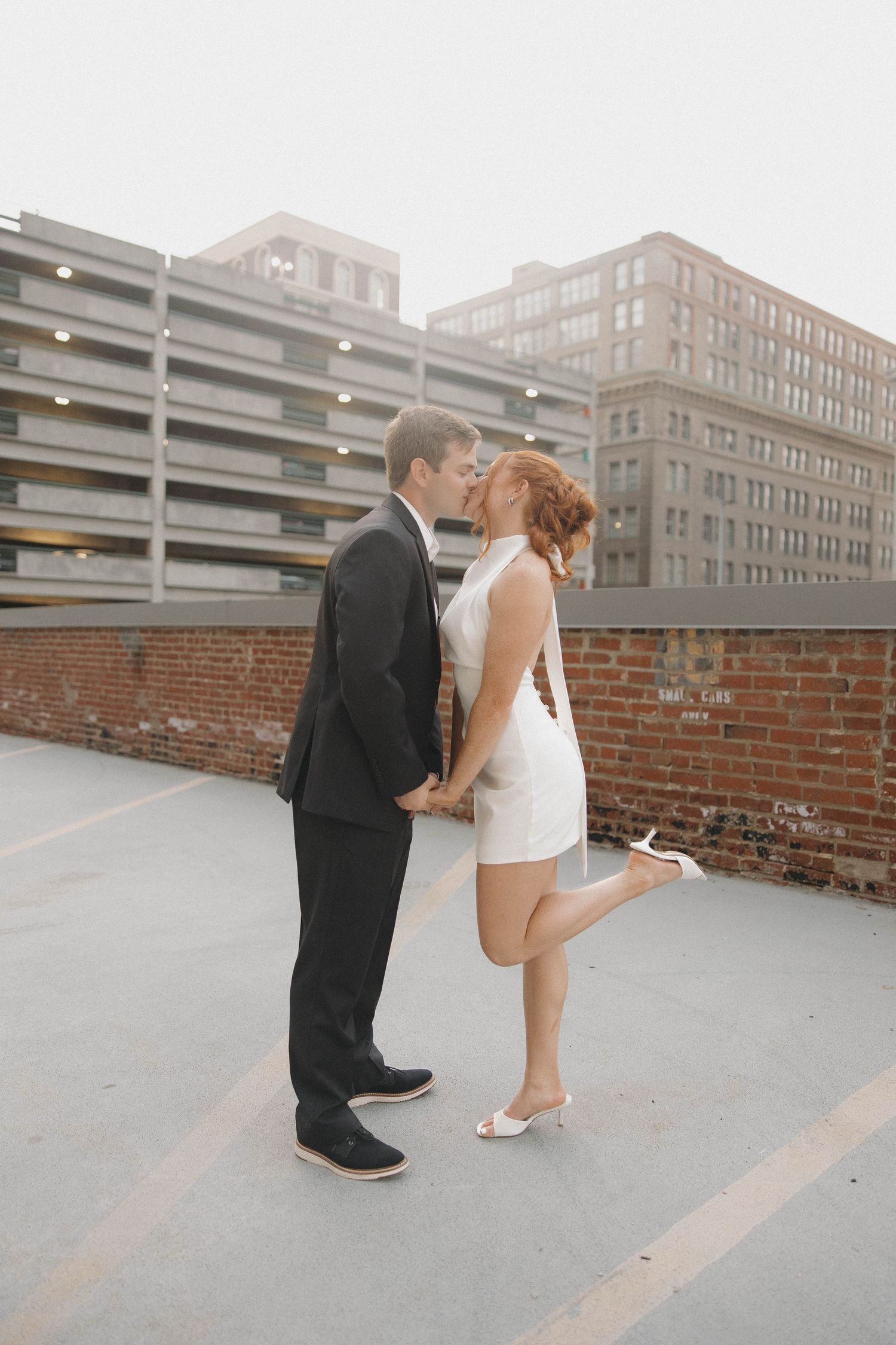 A couple in wedding attire kisses on a rooftop parking garage at dusk with city buildings in the background.