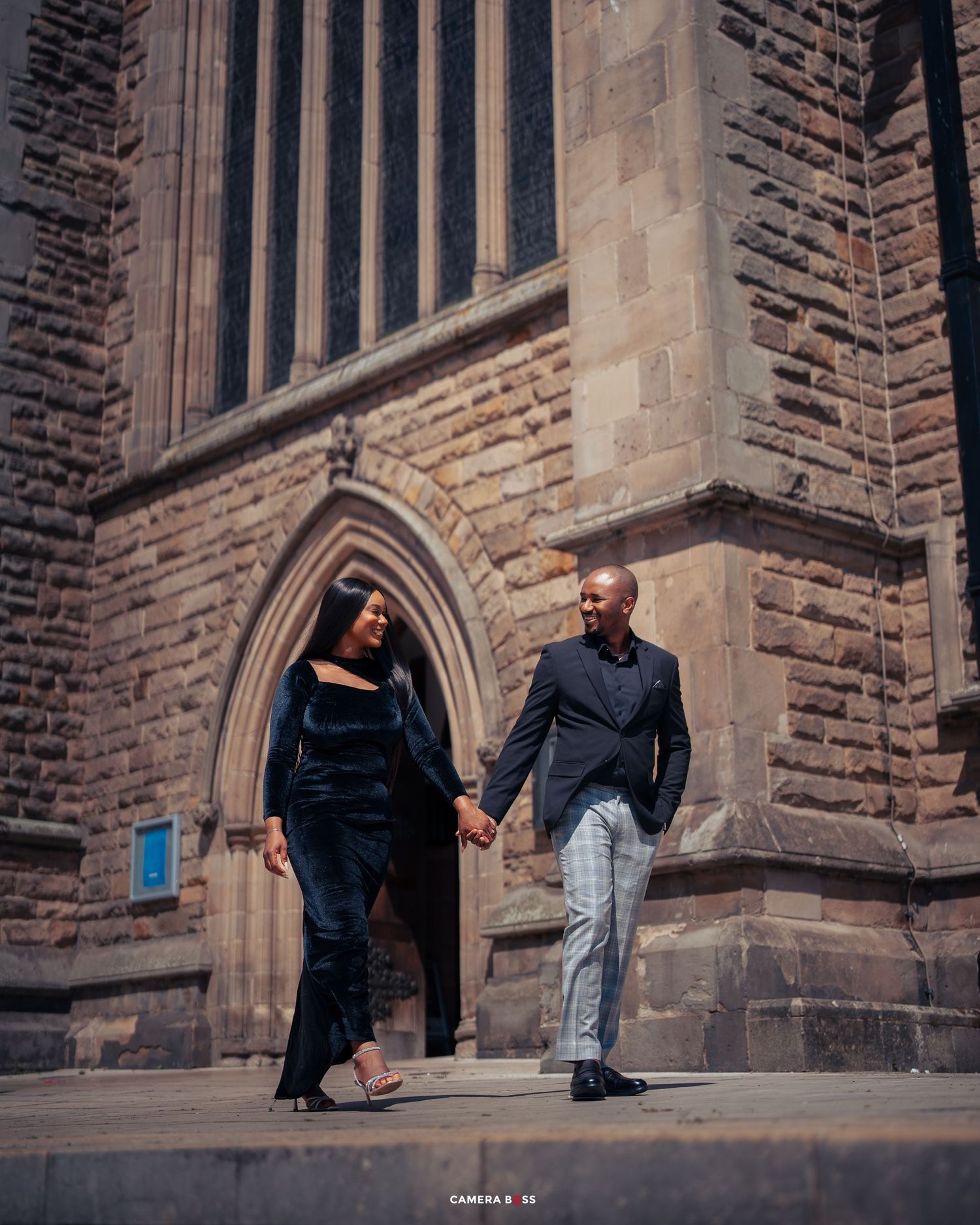 A couple holding hands walks together outside a Gothic church's stone archway at sunset.