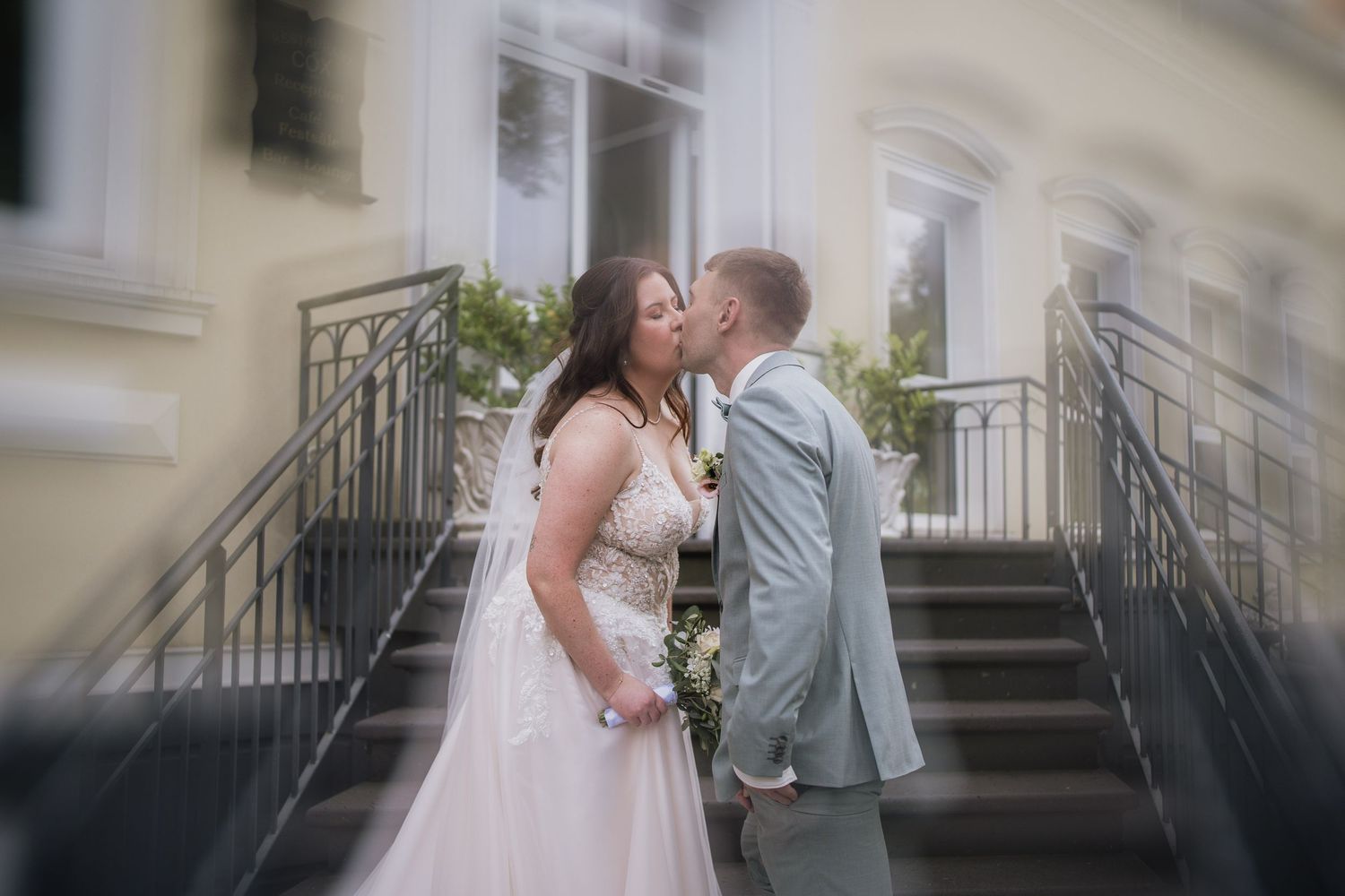 Romantic wedding couple sharing a tender kiss on elegant stone steps with wrought iron railings.