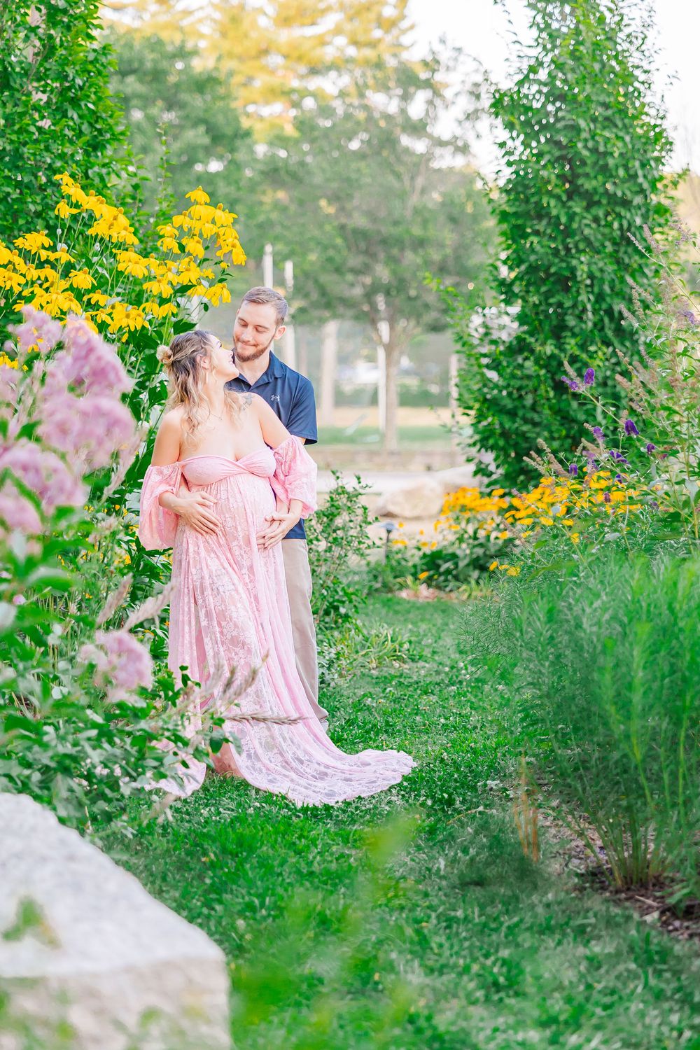 A couple stands together in a lush garden with pink and yellow flowers blooming around them on a summer evening.