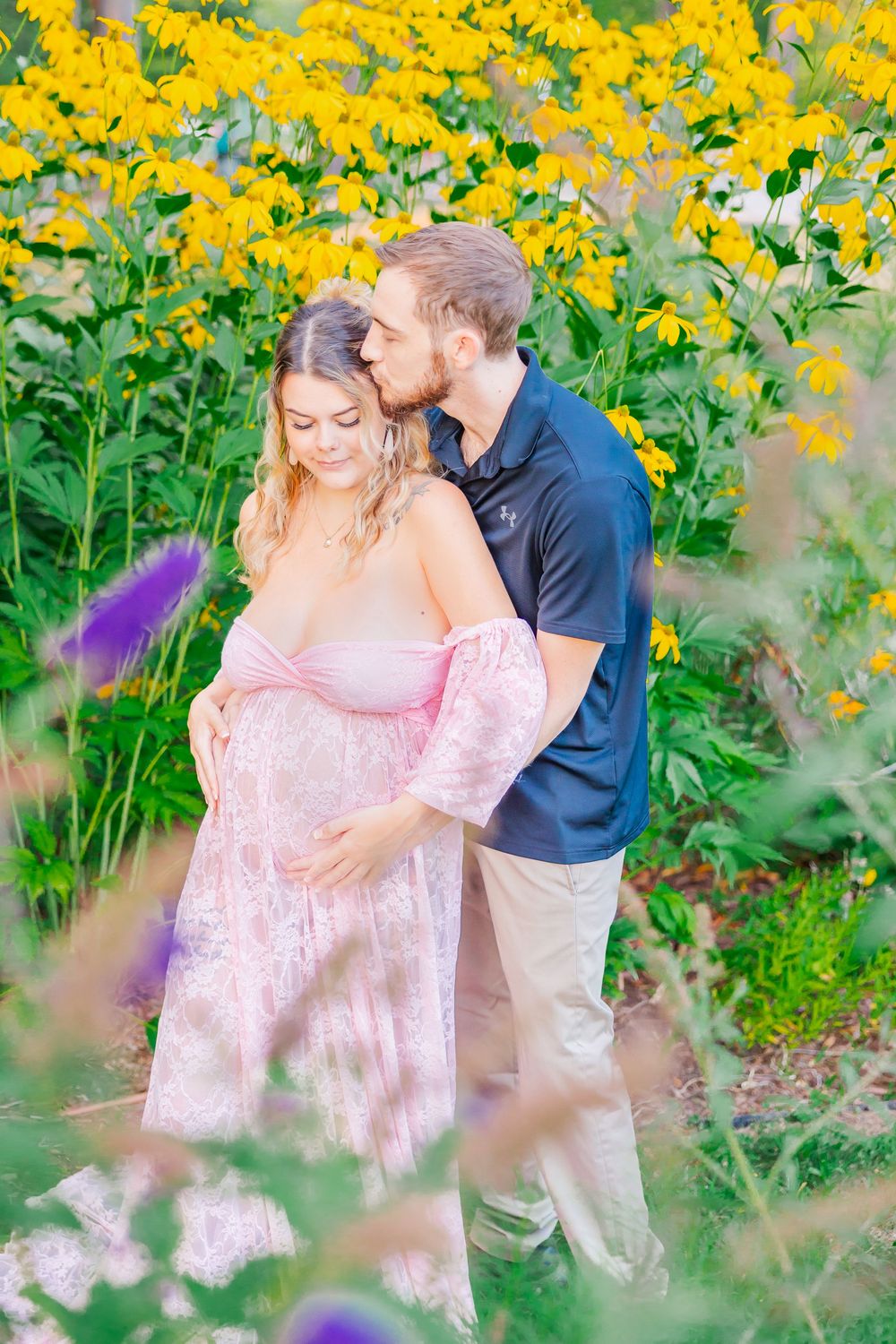 A couple shares a tender moment among bright yellow flowers in a summer garden, with her wearing a pink off-shoulder dress.