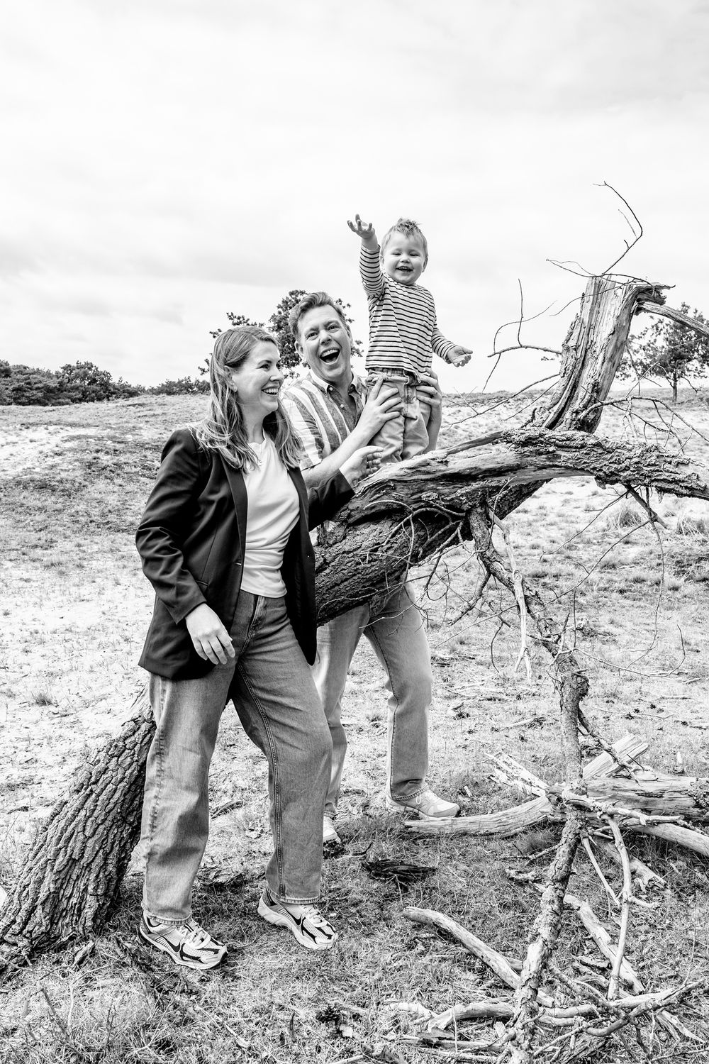 Peuter zwaait naar de camera vanaf een boomstronk terwijl zijn ouders hem vasthouden tijdens een gezinsshoot