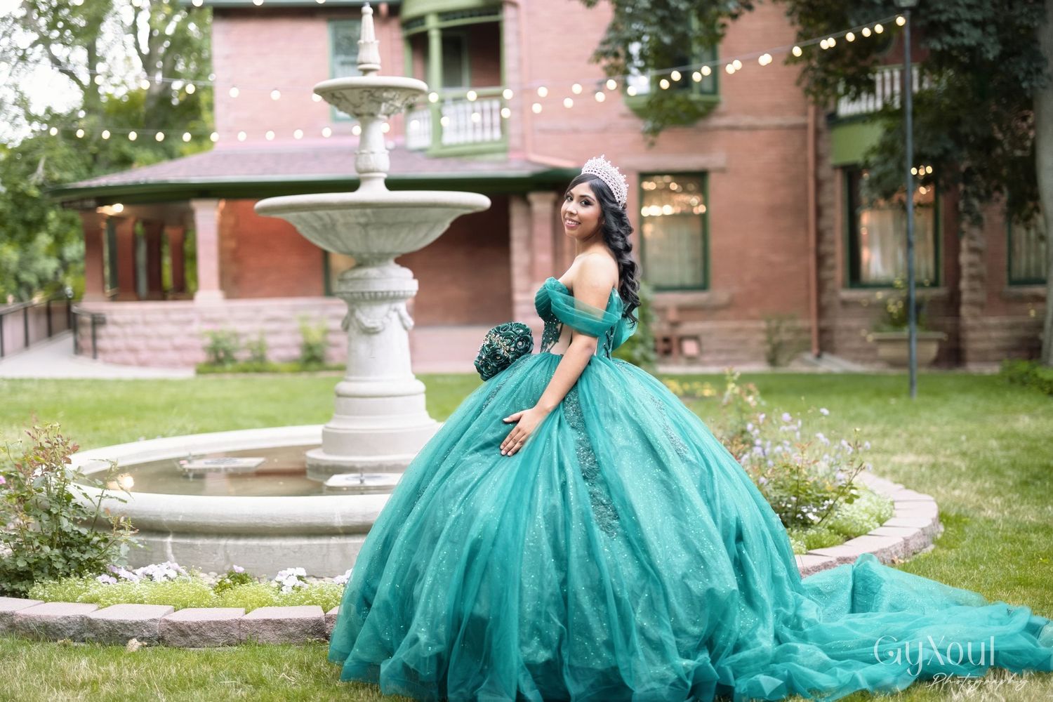 A person in an elegant teal ball gown poses gracefully in a garden setting with a decorative fountain.
