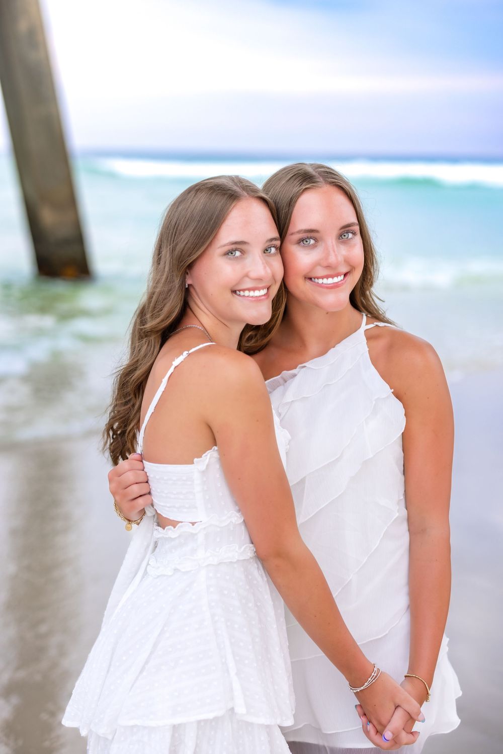 Two women in white dresses pose together on a sunny beach with ocean waves in the background.