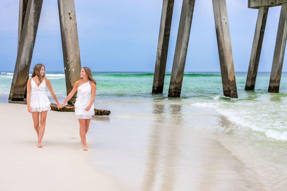 Two people in white dresses stroll along a tropical beach with turquoise water and wooden pier pilings in the background.
