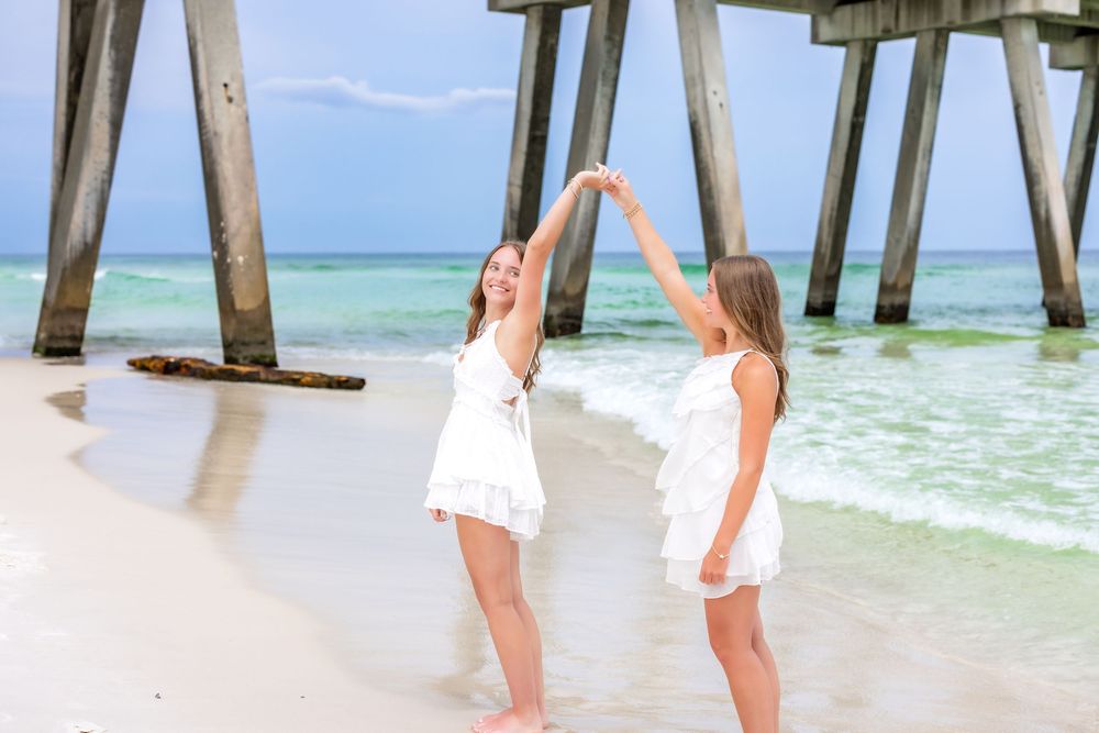 Two people in white dresses walk along a pristine beach with crystal clear waters and wooden pier posts.