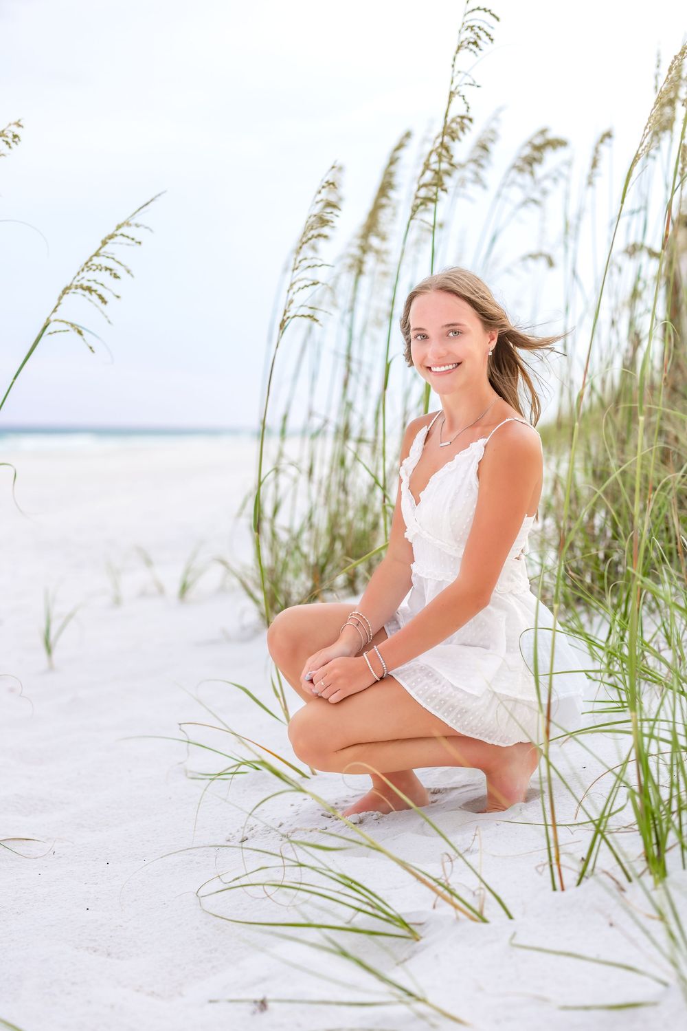 Woman in white dress kneels by sea oats on pristine white sandy beach during summer photoshoot.