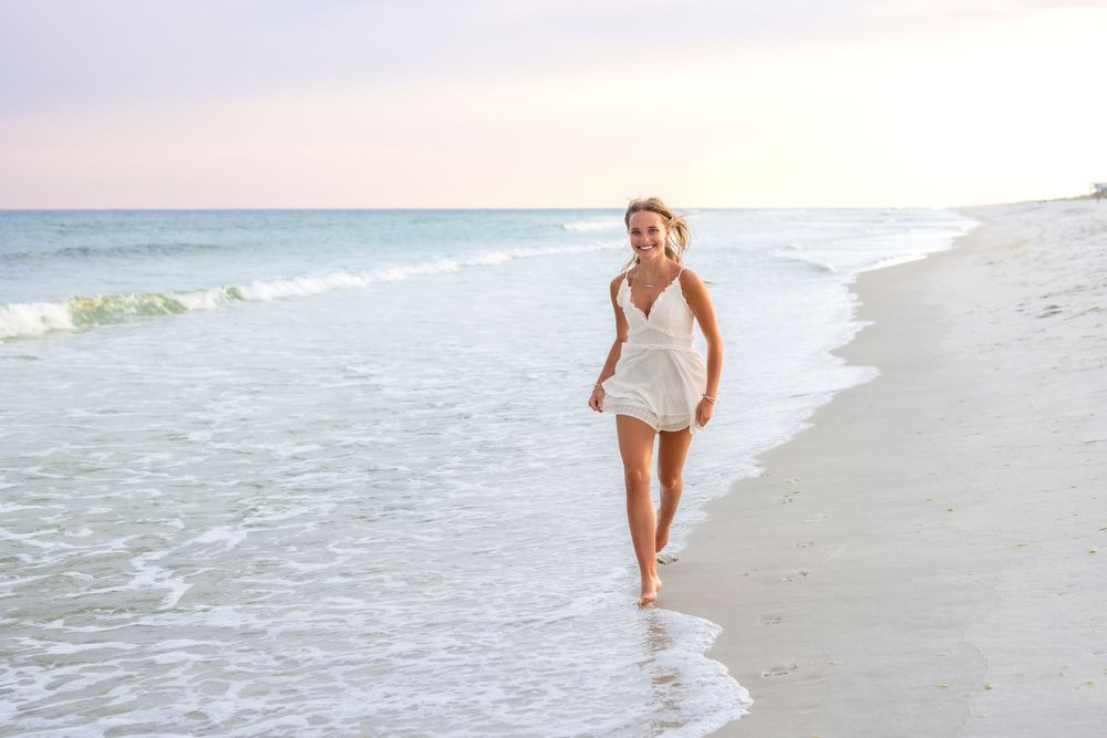 Woman in white dress walks along shoreline at sunset with waves gently rolling in.