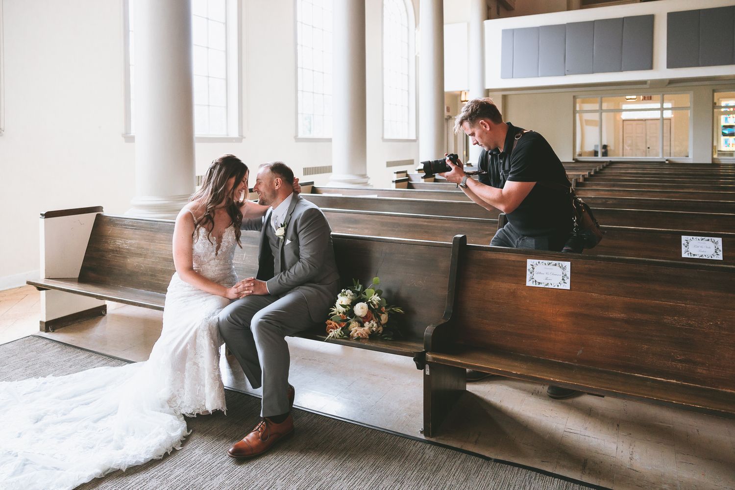 A wedding couple shares an intimate moment on wooden church pews in a light-filled sanctuary.