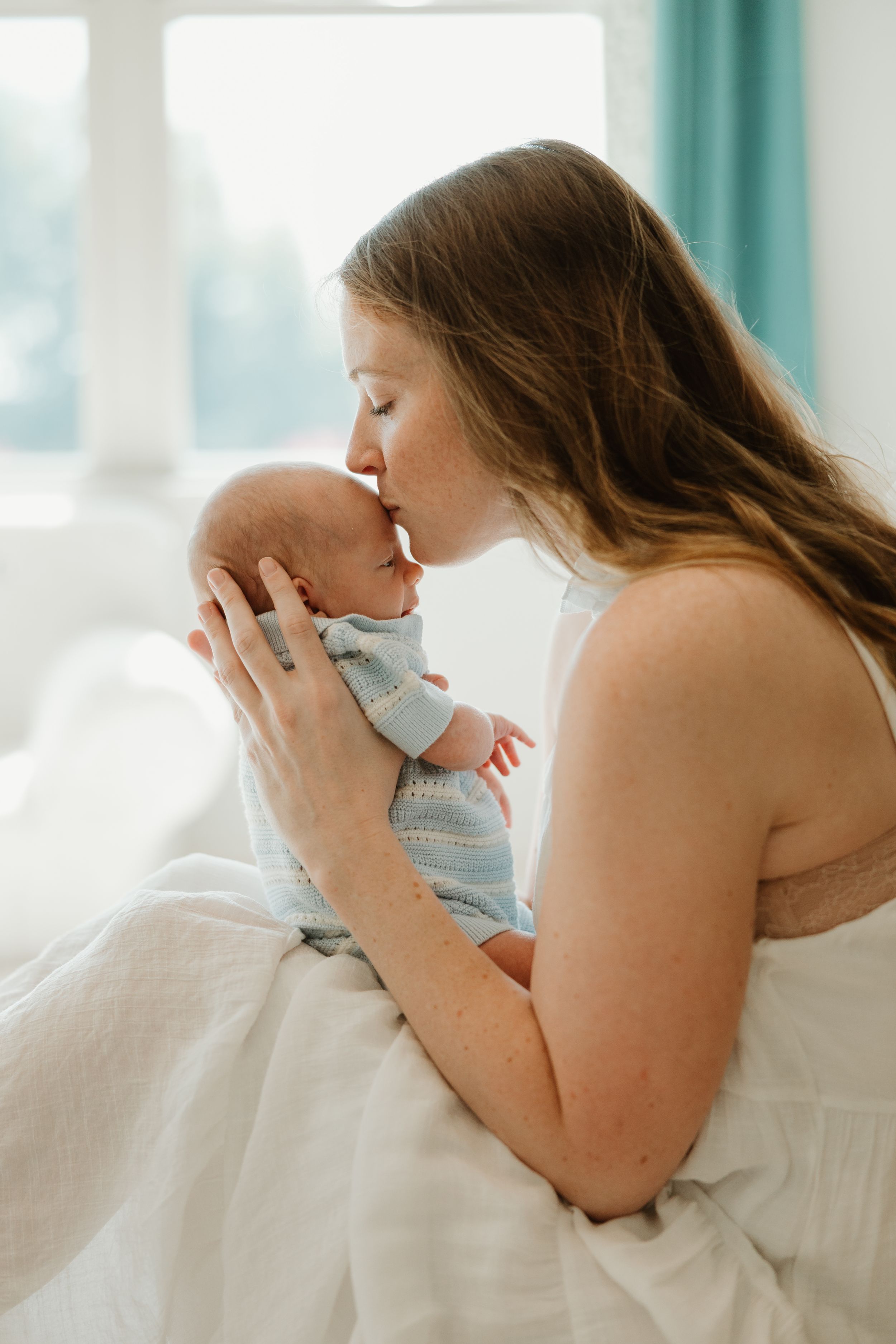 A tender moment captured showing a parent gently kissing their newborn baby while sitting on white bedding.