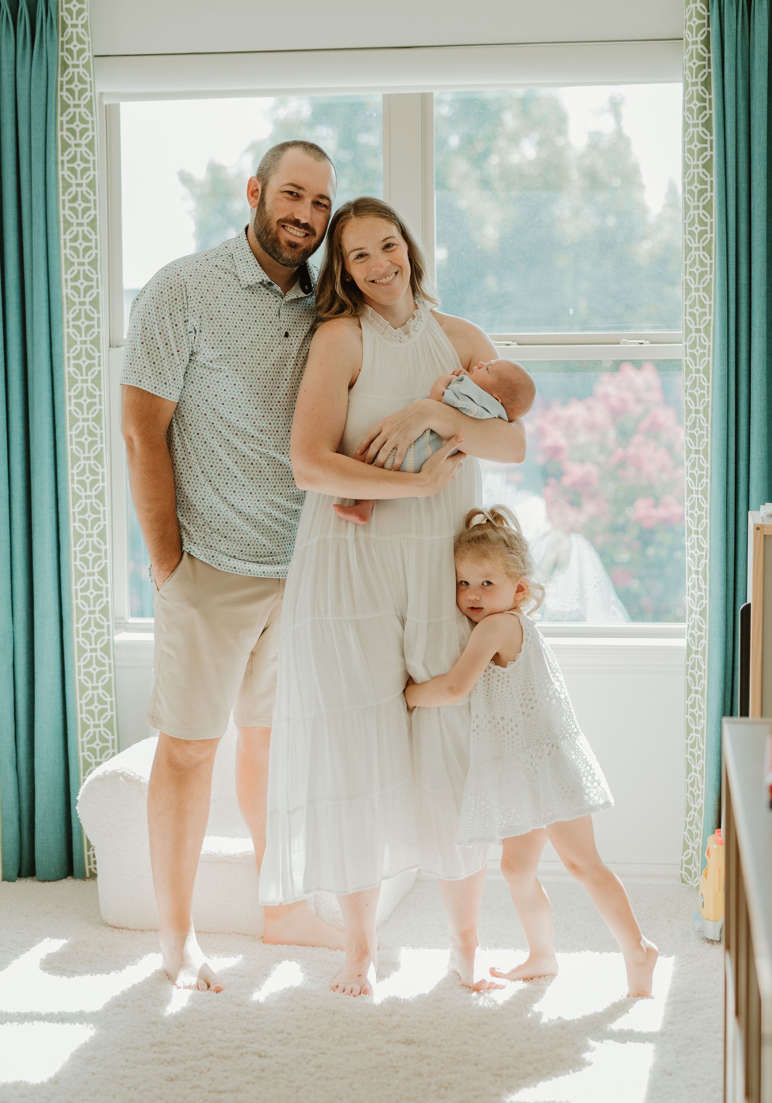 A family wearing light colors poses together in a bright sunlit room with turquoise curtains and hardwood floors.