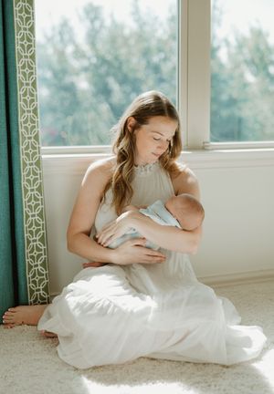 A woman in a white dress sits by a window in soft natural light while nursing a baby.