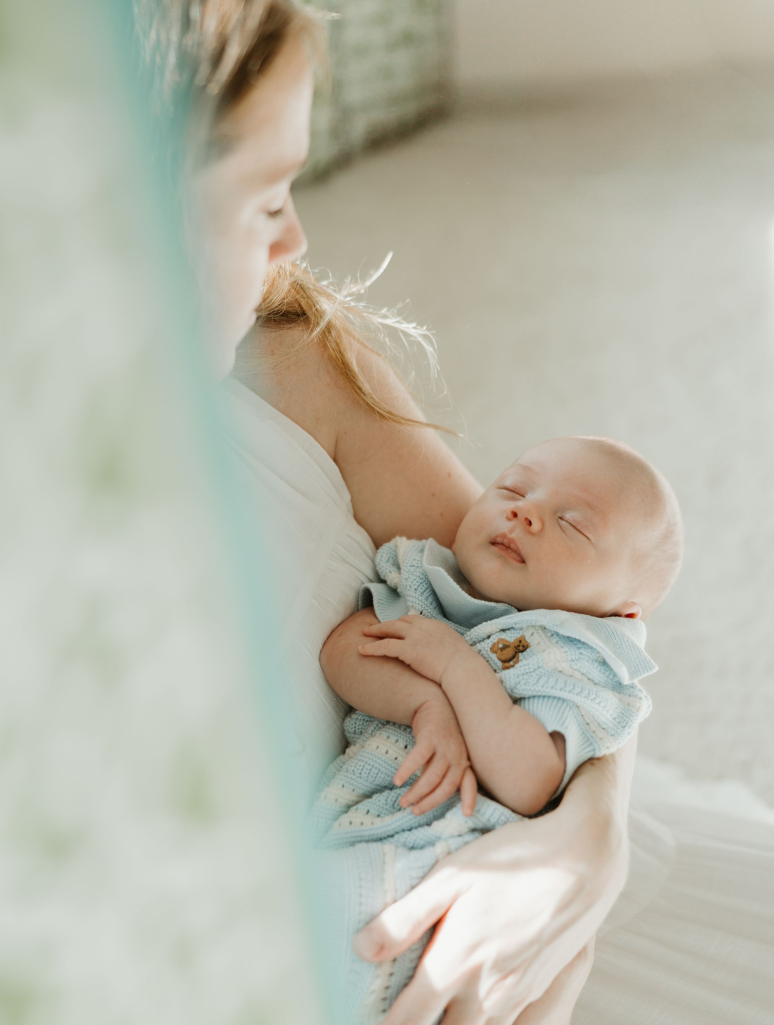 A sleeping newborn in blue clothing rests peacefully against a caregiver in soft natural light.