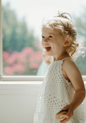 A joyful child wearing a white eyelet dress laughs with pure delight against a soft, blurred background.