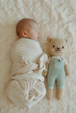 A sleeping baby wrapped in white swaddle lying next to a knitted teddy bear wearing mint green overalls.