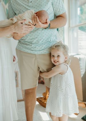 A child in a white dress hugs an adult wearing a mint green shirt and khaki shorts in a sunlit room.