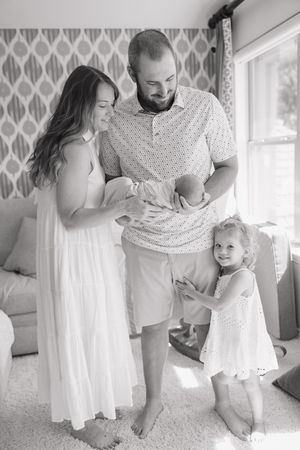 A black and white indoor family photo showing parents with a newborn child and a toddler in matching white outfits.