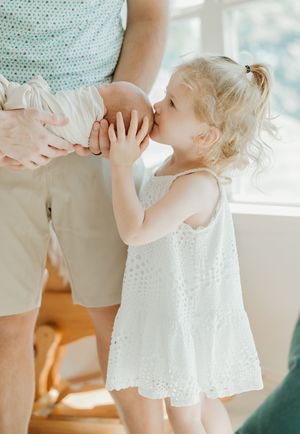 A toddler in a white eyelet dress kisses a pregnant belly in a softly lit room.