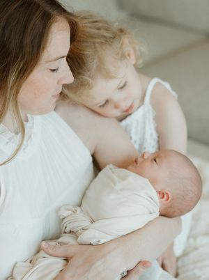 Two young siblings in white clothing share tender moments with their newborn baby sibling against a light background.