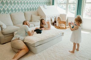 Family lounges together in a bright sunlit room with teal wallpaper and white carpet beside a wooden rocking horse.