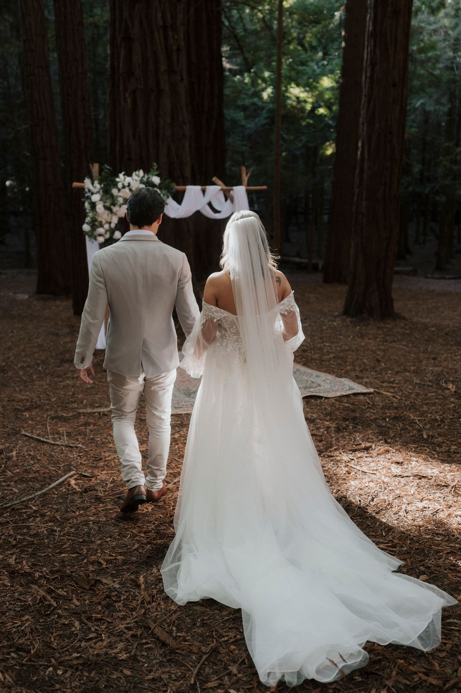 Newlyweds walk together through a redwood forest with bride's flowing white dress trailing behind on pine needles.