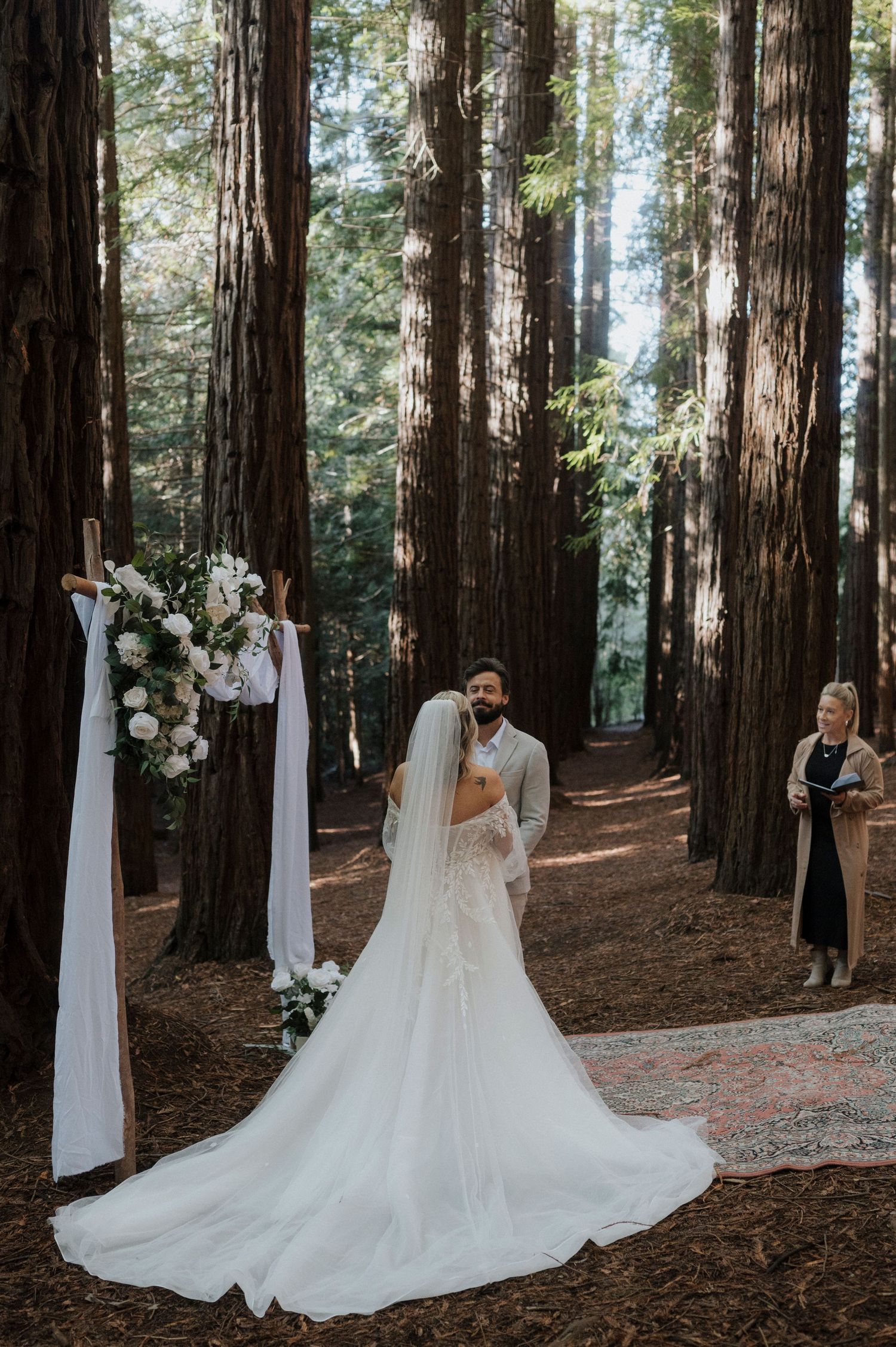 A romantic forest wedding ceremony takes place among towering redwood trees with white fabric draping and floral arrangements.