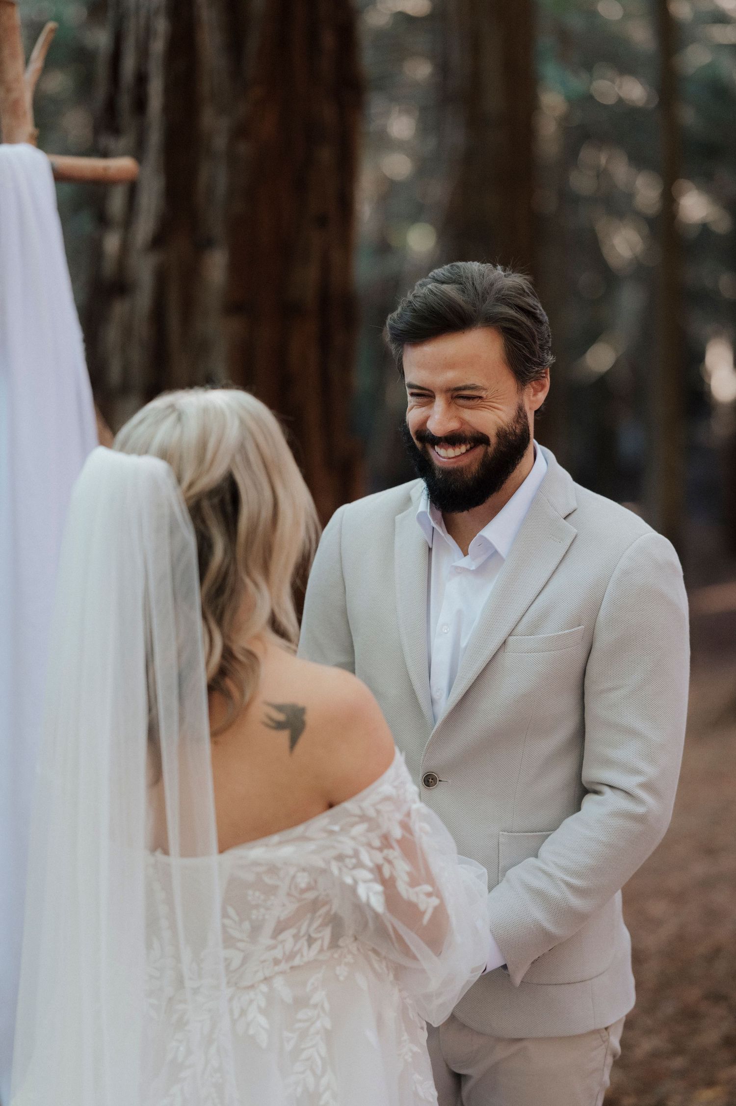 A couple shares a joyful moment during their outdoor wedding in a pine forest setting wearing white formal attire.
